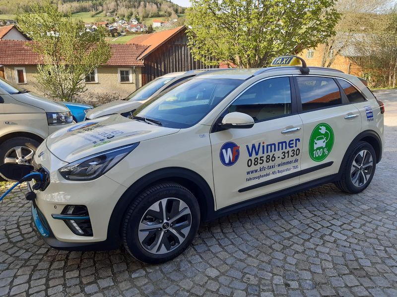 Der cremefarbene Kia e-Niro mit Taxischild auf dem Dach hängt am Ladekabel auf einem gepflasterten Hof. Auf der Fahrertür stehen der Name Wimmer, die Nummer 08586 3130 und eine grüne Plakette mit der Aufschrift 100 Prozent.