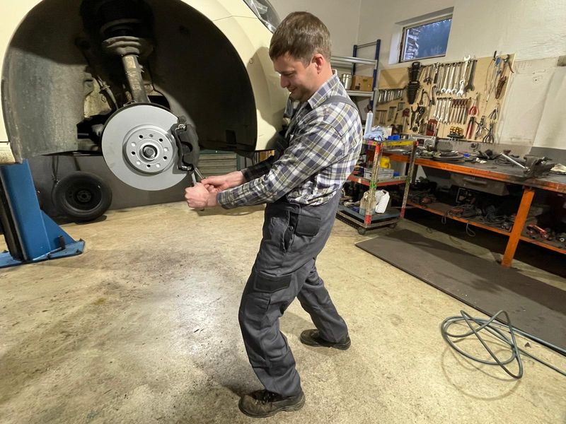 Fritz Wimmer jun. löst an einem aufgebockten Fahrzeug die Bremsscheibe, dahinter die Werkzeugwand der Werkstatt